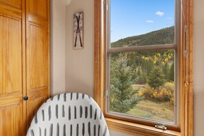 A room with a window overlooking a forested area, a wooden cabinet, and a chair with a striped cushion. - Keystone Getaway at Hidden River Lodge – Balcony Views & Shared Hot Tub (Keystone)