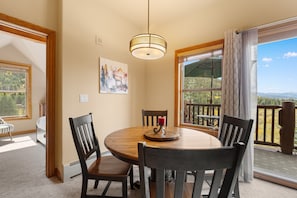 A dining area with a round table, four chairs, a painting, and a view of a mountainous landscape through large windows. - Keystone Getaway at Hidden River Lodge – Balcony Views & Shared Hot Tub (Keystone)