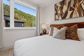 Ein Schlafzimmer mit einem großen Bett, einem Nachttisch mit Lampe, einem Fenster mit Blick auf einen bewaldeten Hügel und einem Bild an der Wand.