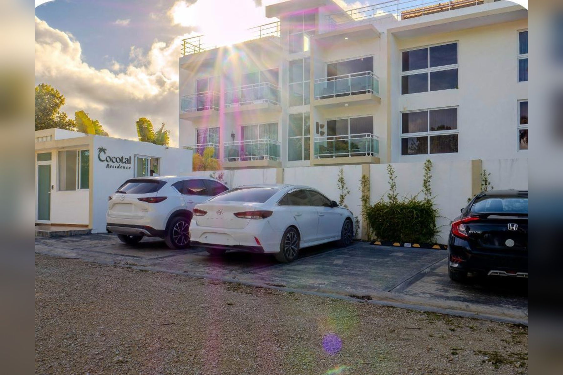 En bygning med flere etager, med et skilt for 'Cocotal Residence'. To hvide biler er parkeret foran.
