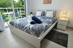 Un dormitorio con una cama, una mesita de noche, una lámpara y vistas al exterior con vegetación.