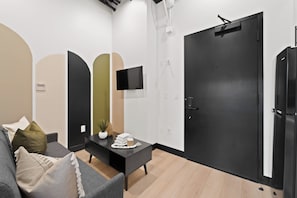 Ein modernes Wohnzimmer mit einer Couch, einem Couchtisch und einem flachbildfernseher an der Wand. Eine schwarze Tür und ein kleiner Topf mit Pflanze stehen auf dem Tisch.