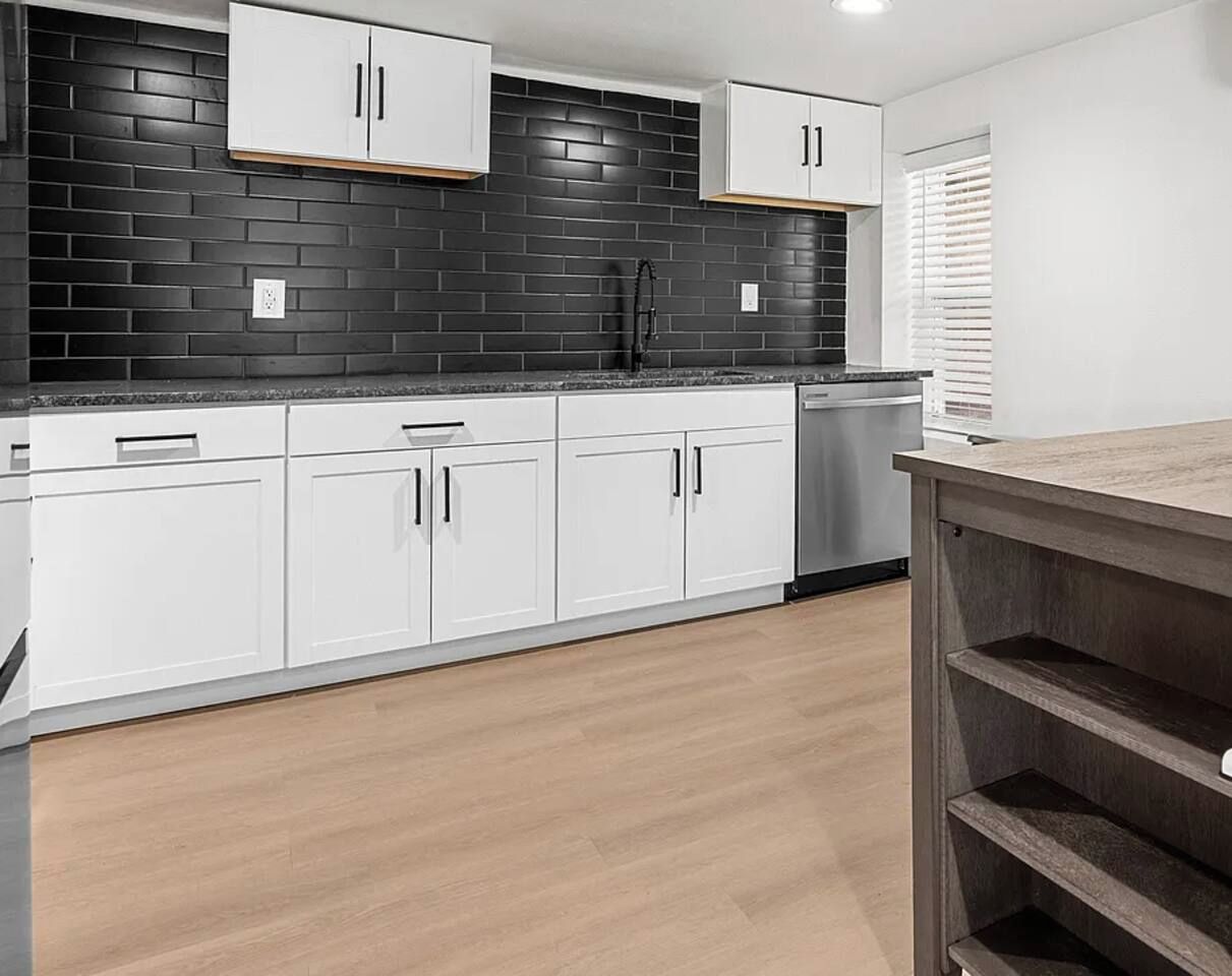 A kitchen with white cabinets, black backsplash, and wooden flooring.