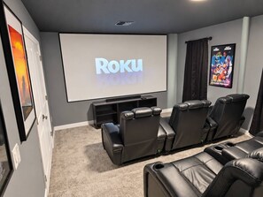 Cine en casa con una pantalla grande mostrando 'Roku', sillones de cuero reclinables, piso alfombrado y carteles de películas enmarcados.