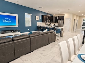 Uma sala de estar moderna com uma grande TV de tela plana, um sofá de couro escuro e uma área de refeições com cadeiras brancas.