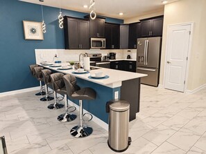 A modern kitchen with dark cabinets, stainless steel appliances, and a central island with bar stools. - Family Paradise | 8BR | Champions Gate Resort (Kissimmee)