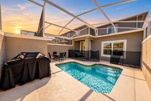 Ein Schwimmbad mit überdachtem Grill und Stühlen, umgeben von einer überdachten Terrasse mit Blick auf den Sonnenuntergang.