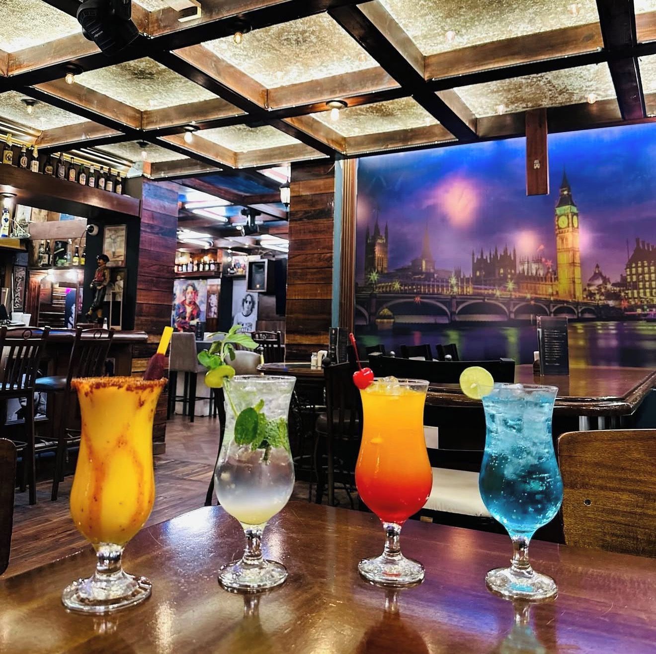 Baarialue, jossa on neljä värikästä cocktail-drinkkiä puupöydällä, taustalla kaupunkimaisemaa esittävä seinämaalaus sekä baaritiski, jossa on erilaisia pulloja ja laseja.