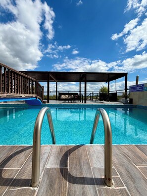 A swimming pool with clear blue water, surrounded by a wooden deck and a covered area with tables and chairs. - Hacienda Real de Linares (Linares)