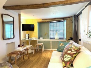 A cozy room with a wooden bench, a sofa, a TV, a small dining table, and a window with blinds. - The Coach House, Market Deeping by Belmont Places (Market Deeping)