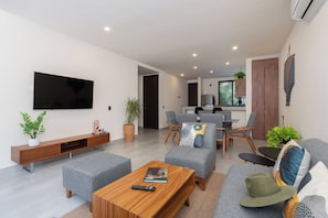 A modern living room with a flat-screen TV, grey sofas, a wooden coffee table, and a remote control on it. - Resort-Style 2-Bedroom | Padel, Pool, Grill, Coworking Space (Tulum)