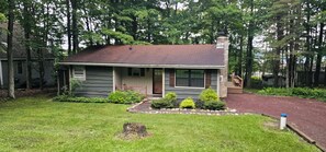 A single-story house with a front porch, a chimney, and a small garden area with shrubs. - Cozy 3 BR Cottage-Walk to Lake-Minutes to Skiing-Make Lasting Memories (Pocono Lake)