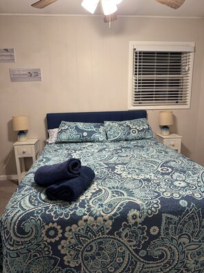 A neatly made bed with a patterned bedspread, two bedside tables with lamps, a window with blinds, and a ceiling fan. - Cozy 3 BR Cottage-Walk to Lake-Minutes to Skiing-Make Lasting Memories (Pocono Lake)