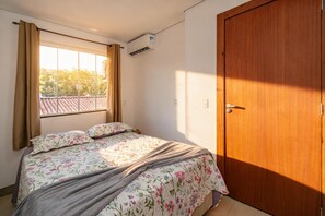 Une chambre à coucher avec un lit, des rideaux, un appareil de climatisation, une porte en bois et une fenêtre donnant sur un toit recouvert de tuiles.