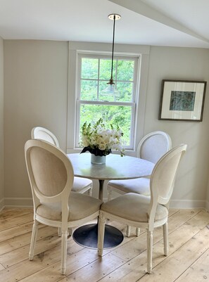 A dining area with a round table, four chairs, a vase of flowers, a hanging light fixture, and a window with a view of greenery. - The Flirt - Restored 1927 Cottage at Lake Waramaug (New Preston)