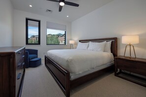 Ein Schlafzimmer mit einem großen Bett, einem Schreibtisch, einem Stuhl, einem Nachttisch mit Lampe und zwei Fenstern mit Jalousien.