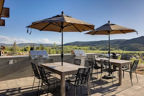 Eine Terrasse im Freien mit Tisch und Stühlen, zwei Grillstationen und Sonnenschirmen, die Schatten spenden.