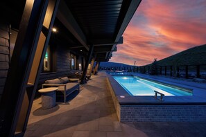 Ein Poolbereich im Freien mit Blick auf den Sonnenuntergang, eine überdachte Lounge mit Sofas und Tischen sowie eine Backsteinmauer.