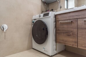 A compact washing machine in a bathroom with a wooden cabinet and a towel rack. - Luxurious Panoramic Apartment (Tiranë)