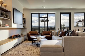 Ein modernes Wohnzimmer mit einer Couch, Sesseln, einem Couchtisch und einem Kamin. Große Fenster bieten einen Ausblick ins Freie.