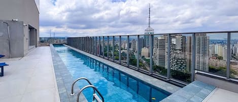 A rooftop swimming pool with a city view, a glass railing, and a clear sky.