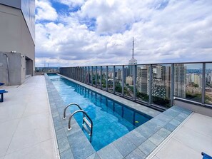 Una piscina en la azotea con vistas a la ciudad, barandilla de cristal y cielo despejado.