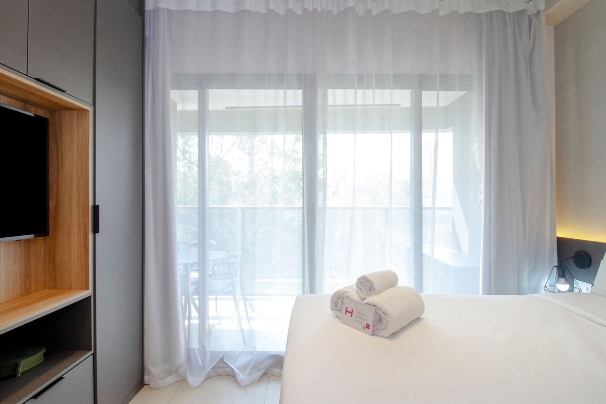 Habitación de hotel con cama, televisor a la izquierda, ventana con cortinas traslúcidas y una toalla sobre la cama.