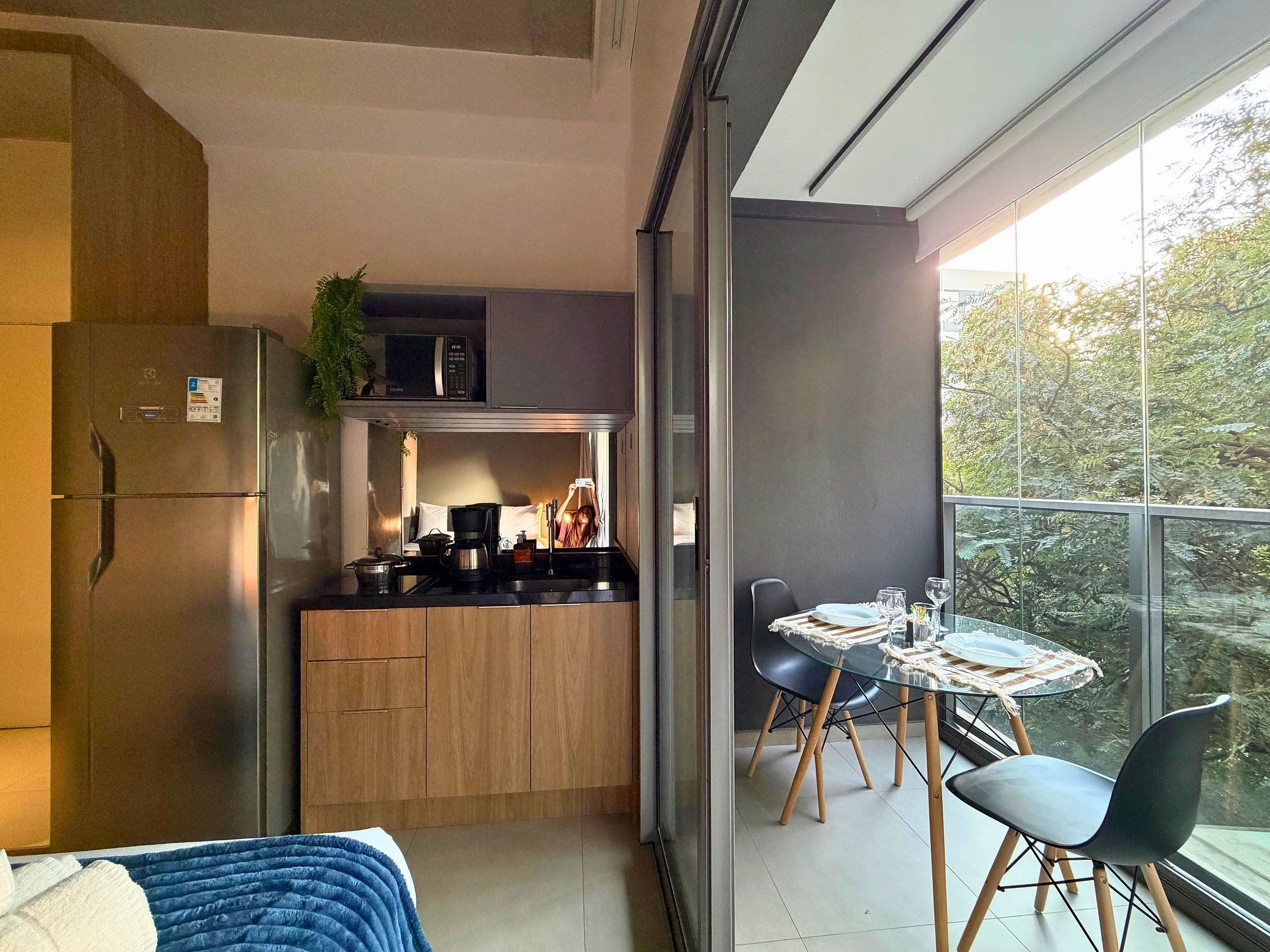 Een compacte keuken met een koelkast, magnetron en kookplaat, een eethoek met een tafel en stoelen, en een balkon met uitzicht op bomen.