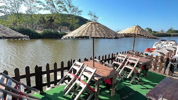 Una terraza a la orilla del río con mobiliario de madera, incluyendo mesas y sillas, y dos sombrillas de paja.
