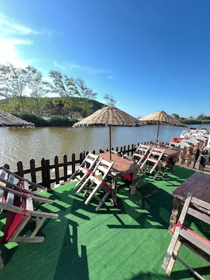 Una terraza a la orilla del río con mobiliario de madera, incluyendo mesas y sillas, y dos sombrillas de paja.