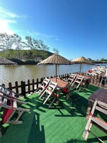 A riverside deck with wooden furniture, including tables and chairs, and two thatched umbrellas.