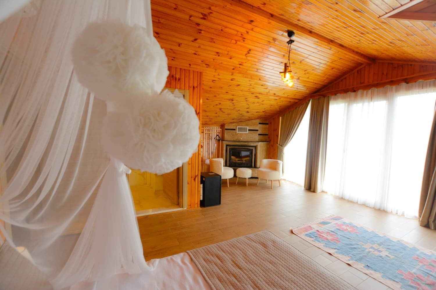 Una habitación acogedora con techo de madera, chimenea, dos sillones y un ventanal con cortinas.