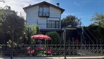 Un bâtiment à deux étages, avec un mur de pierre à l’avant, un parasol rouge et un escalier menant à une zone abritée.