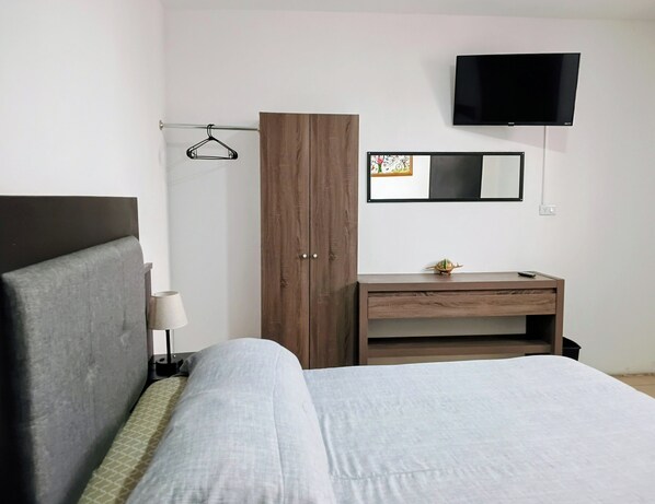 A hotel room with a bed, a wooden wardrobe, a desk with a small turtle figurine, a wall-mounted TV, and a framed picture.