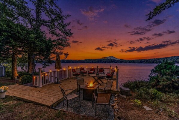 A wooden deck with outdoor seating and a fire pit, overlooking a body of water at sunset.
