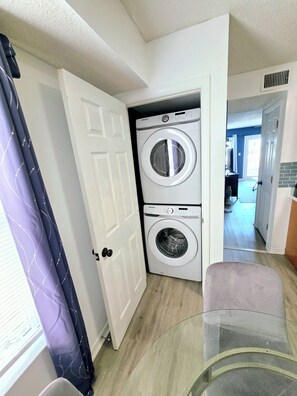 A laundry room with two stacked washers and dryers, a white door, and a view into a living area with a blue wall and a door leading to another room. - 1 Block to Beach | 3BR Gulf Shores Home, Sleeps 8 (Gulf Shores)