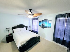 A bedroom with a bed, a ceiling fan, a nightstand, a lamp, and a window with curtains. - Pirate’s Hideaway  3B/2.5B  Sleeps 8 Beach 1 Block (Gulf Shores)