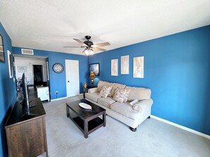 A living room with a blue wall, a ceiling fan, a beige sofa, a coffee table, and a television on a wooden stand. - Pirate’s Hideaway  3B/2.5B  Sleeps 8 Beach 1 Block (Gulf Shores)