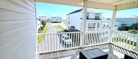 A covered outdoor deck with a wooden floor, a seating area with a sofa and a table, and a view of a residential street with houses and a clear sky.