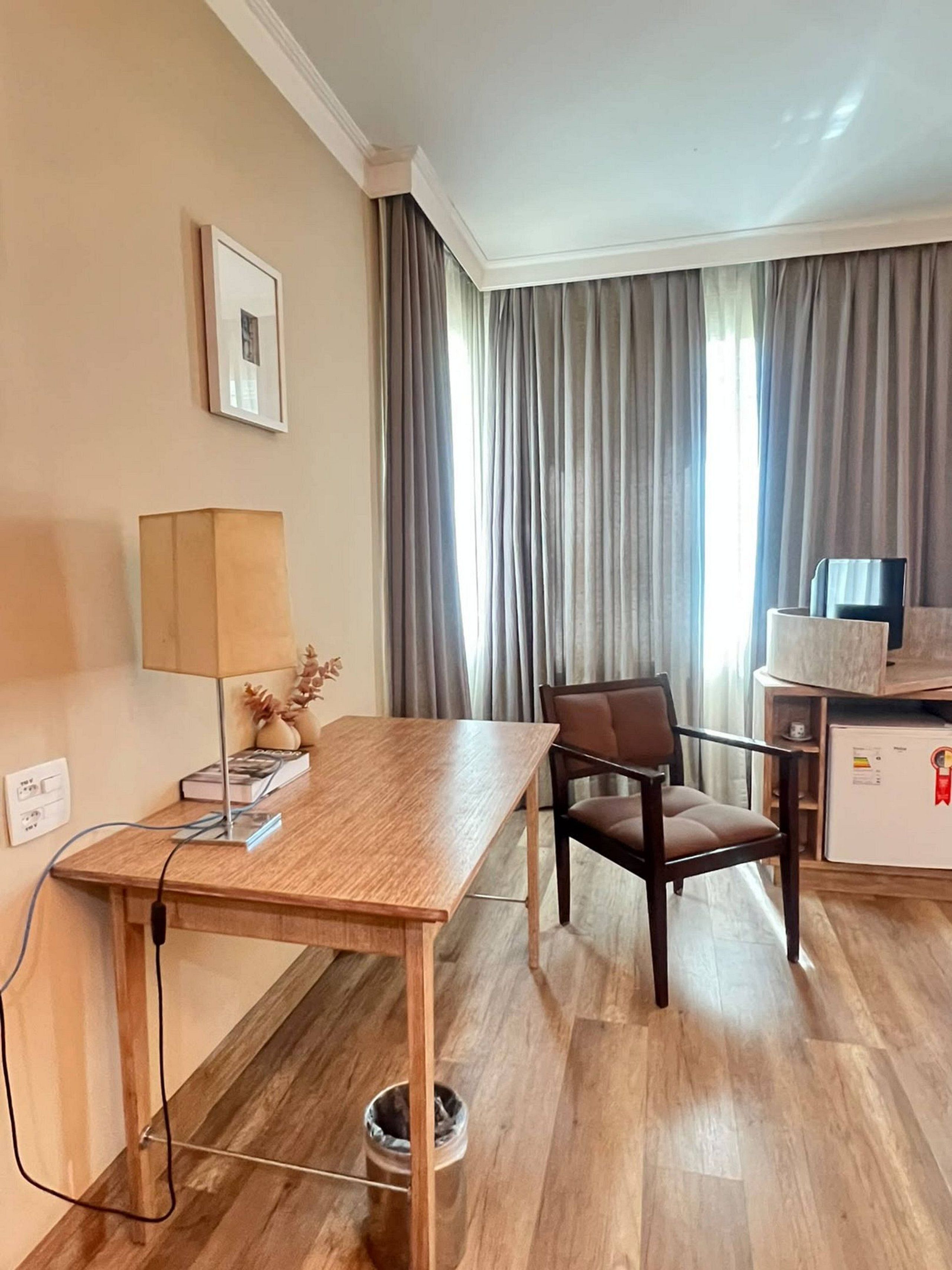 Habitación de hotel con un escritorio de madera, una silla, una lámpara, un cuadro en la pared y una ventana con cortinas.