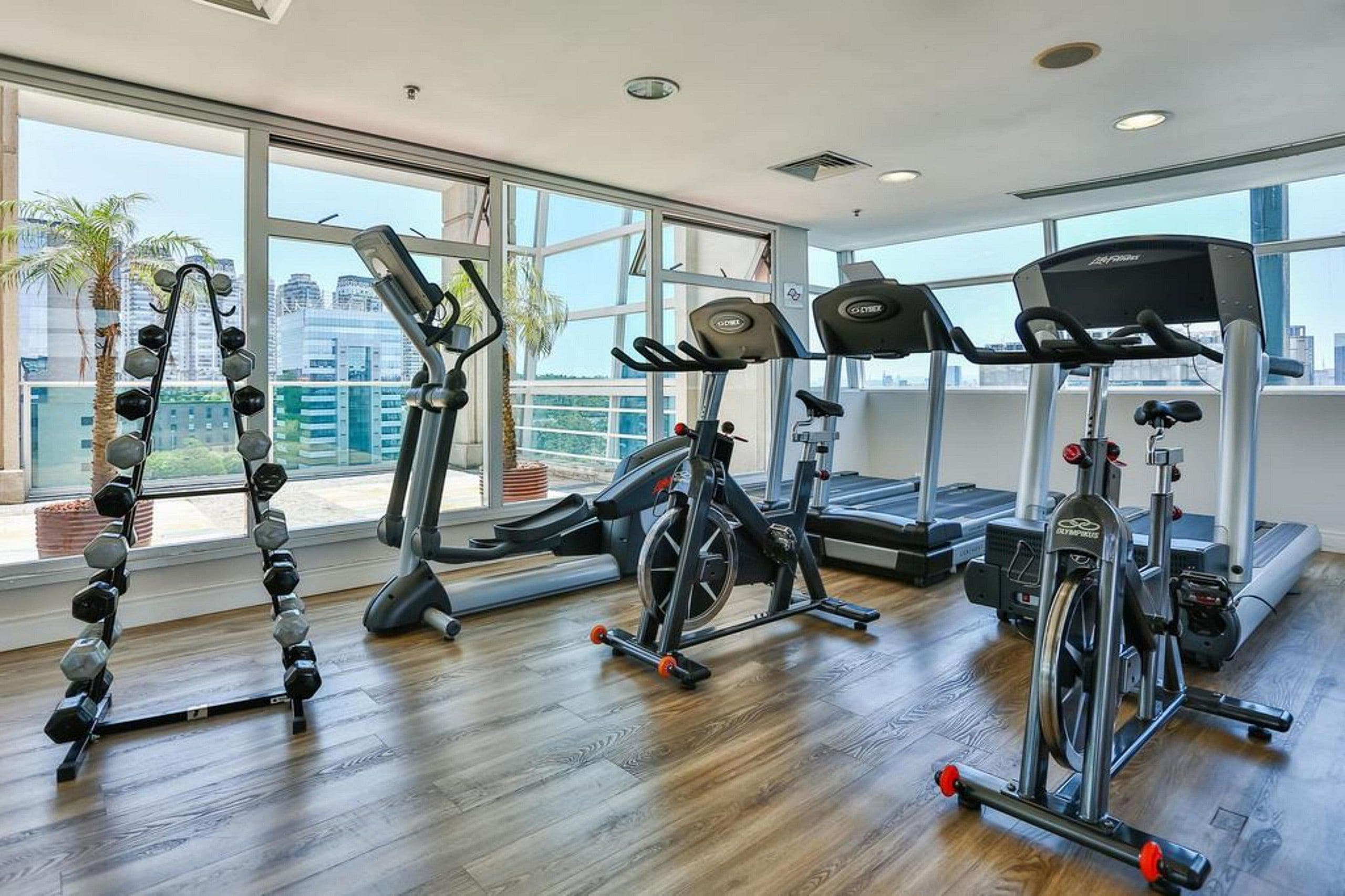 Un gimnasio con diversos equipos de ejercicio, ventanas amplias y vistas a la ciudad.