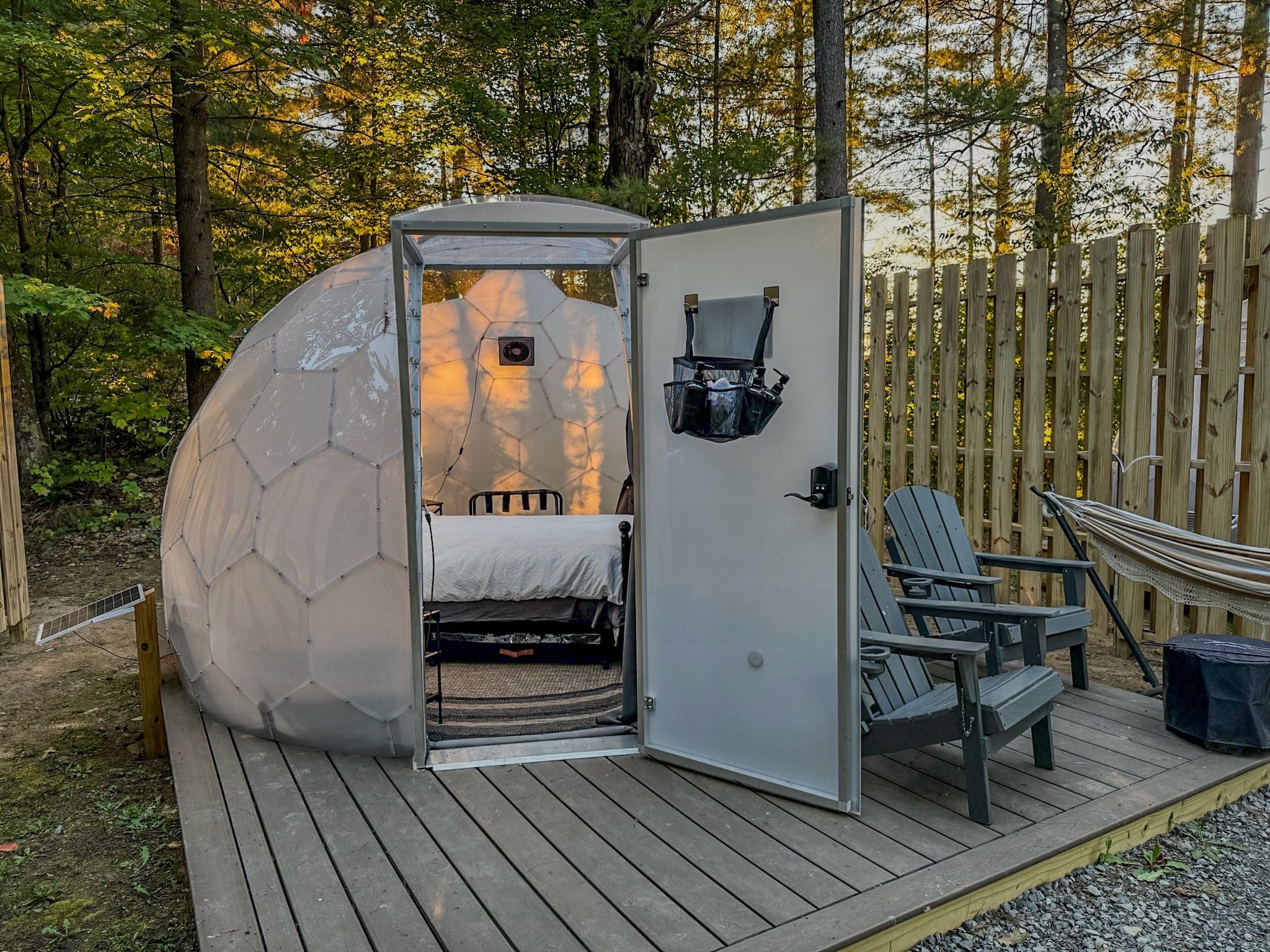 A geodesic dome structure with a bed and seating area, surrounded by a wooden deck and a wooden fence.