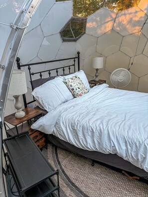A bed with white bedding, a lamp, a fan, and a small table in a room with a geometric patterned wall. - Capricorn | Black Lake Glamping Dome (Hammond)