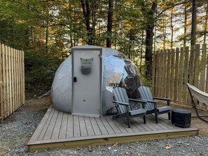 A portable sauna with a door, two Adirondack chairs, and a hammock on a wooden deck surrounded by trees. - Capricorn | Black Lake Glamping Dome (Hammond)
