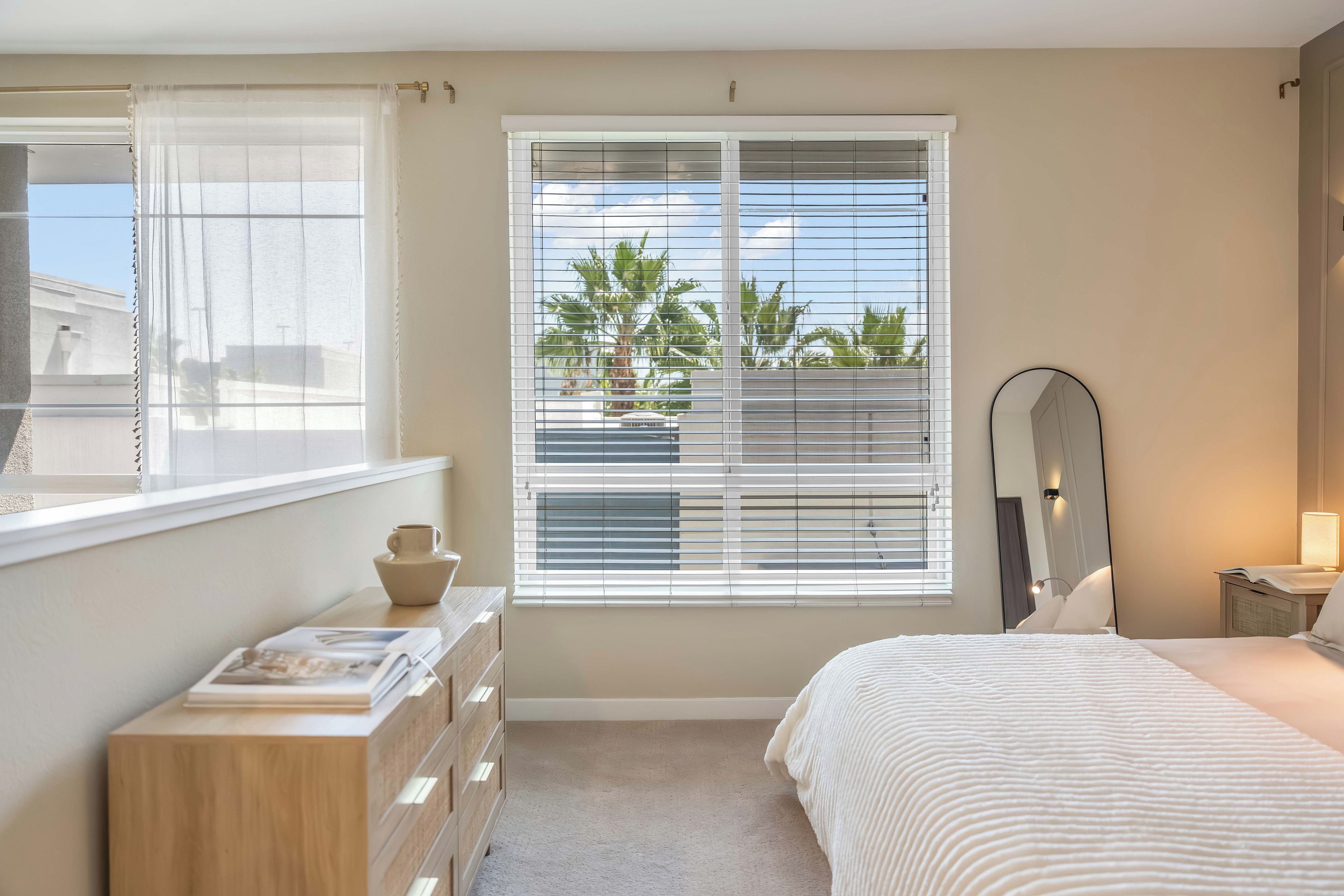 A bedroom with a bed, a dresser, a window with blinds, a mirror, and a bedside lamp.