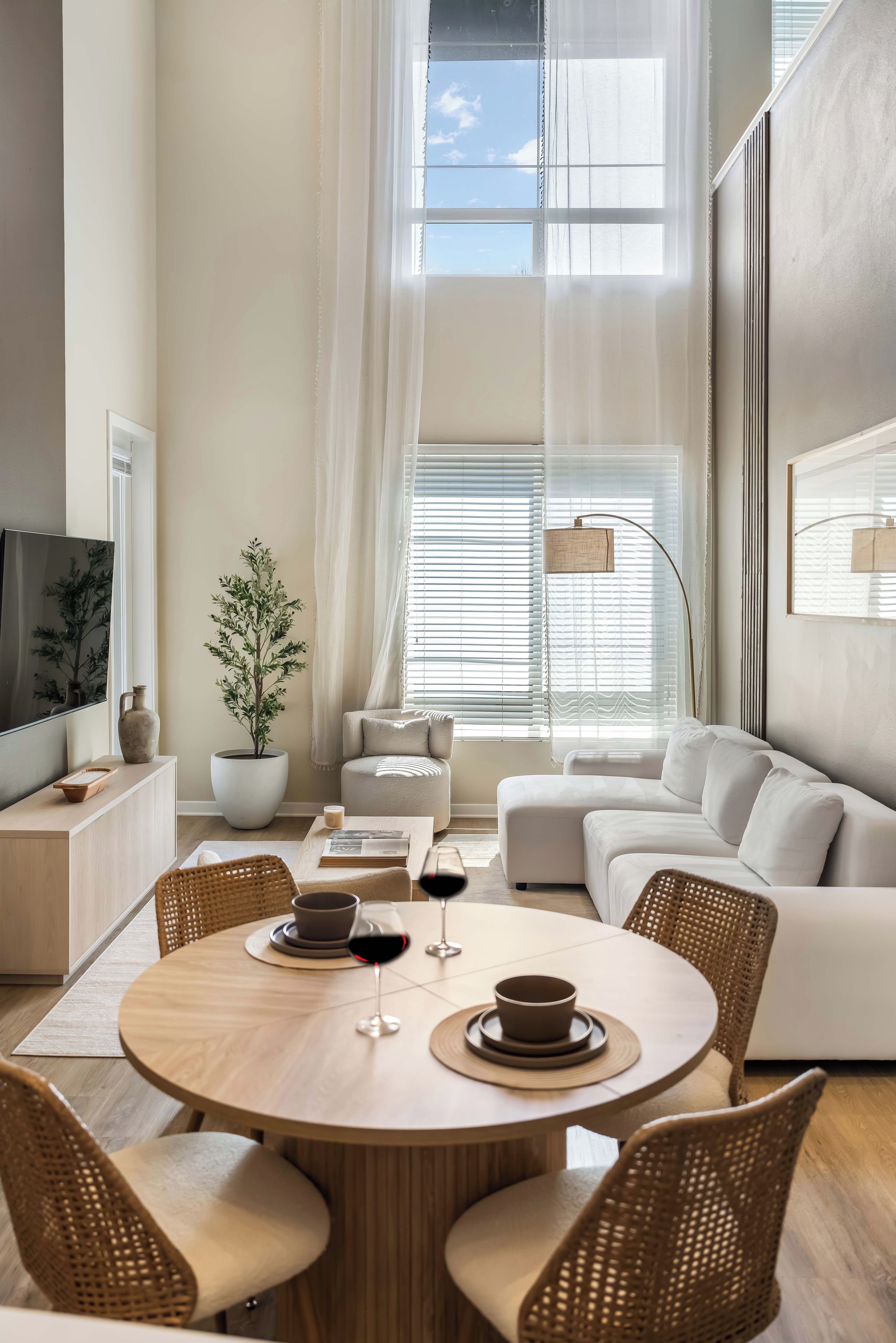 A modern living room with a round wooden table, white sofas, and a large window with blinds.