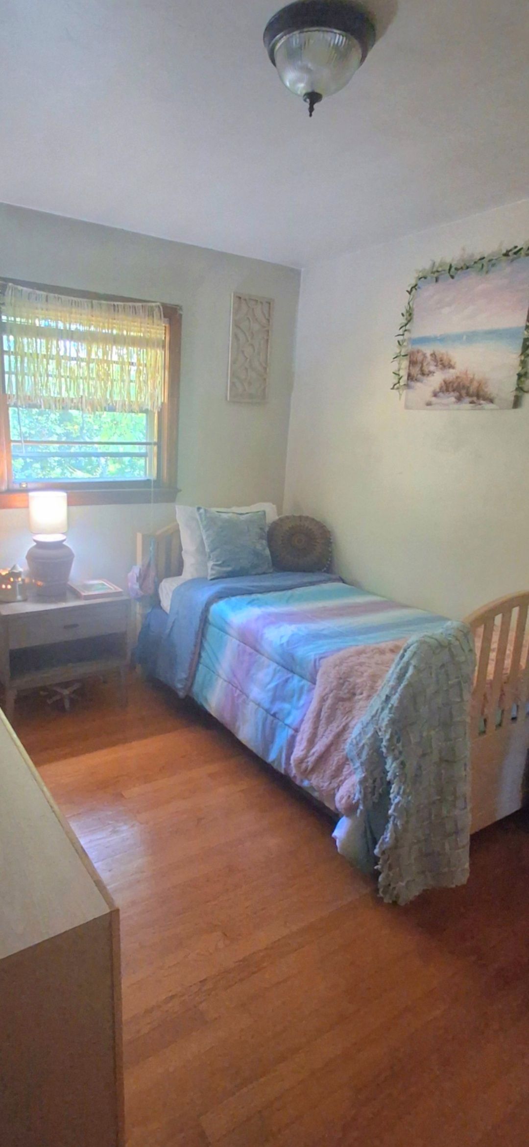 A bedroom with a bed, a nightstand, a lamp, a window with curtains, and a wall-mounted painting.
