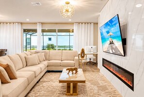 A modern living room with a sectional sofa, a fireplace, a mounted TV, and a view of the outdoors through large windows. - Amazing 10BR Single Family at Windsor Island 49LA (Davenport)