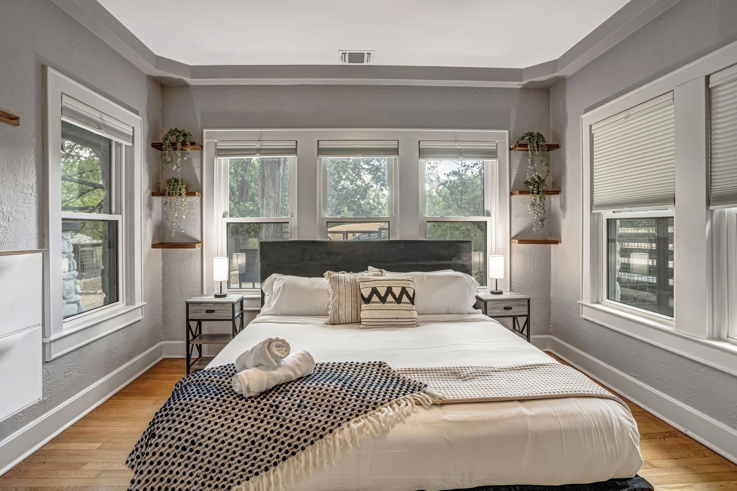 A bedroom with a large bed, two bedside tables, a headboard, and three windows with plants.