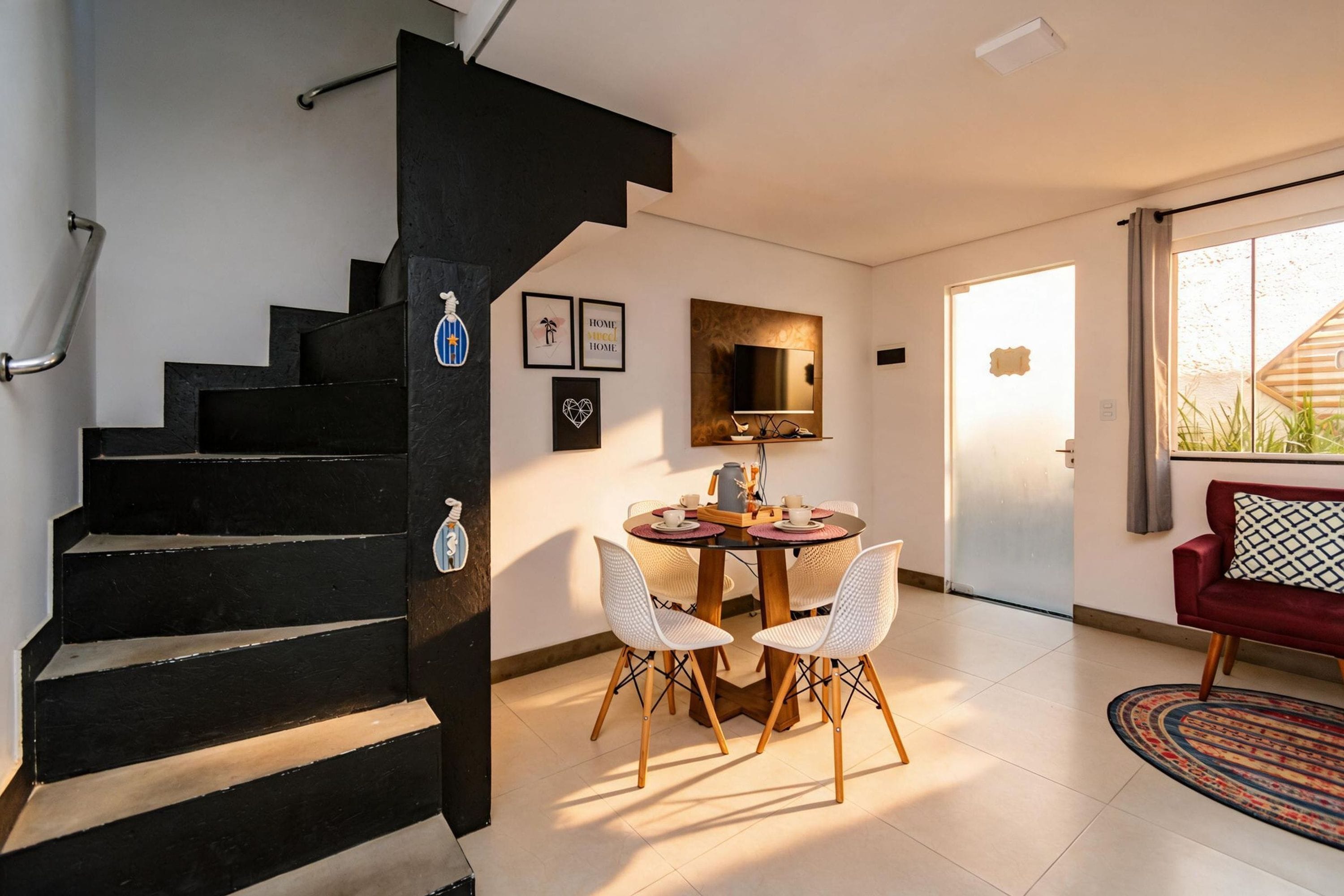 Een trap leidt naar een eethoek met een tafel en stoelen, een aan de muur bevestigde televisie en een rode bank in een lichte kamer met uitzicht door een glazen deur.
